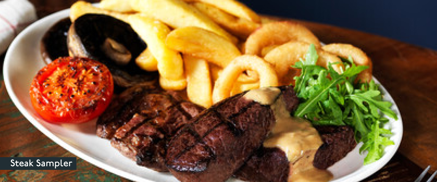 A Beefeater Steak Sampler with chips, mushrooms, a tomato, onion rings, rocket and a selection of steak on a white oval plate.